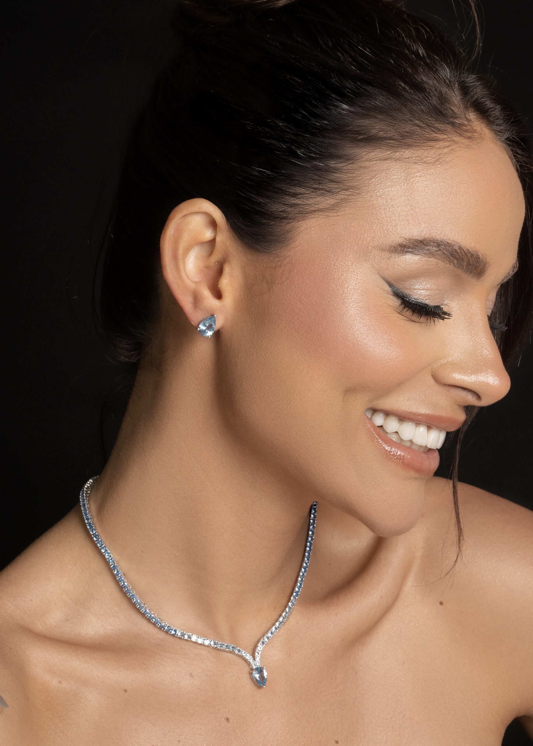 Modelo sorrindo com colar riviera prateado e brinco de pedra azul